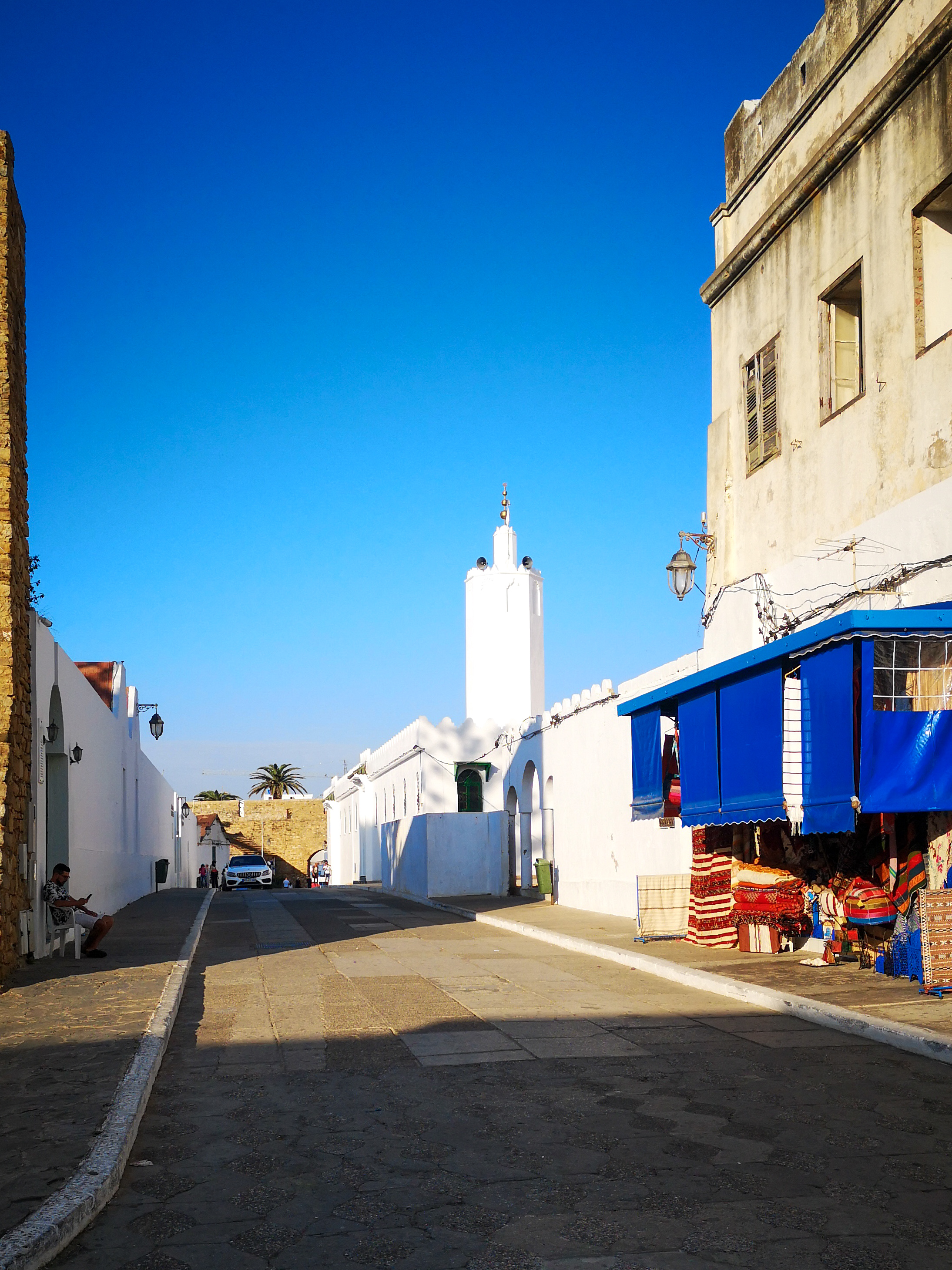 Rue d'Asilah avec une mosquée blanche au fond et une boutique à auvent bleu