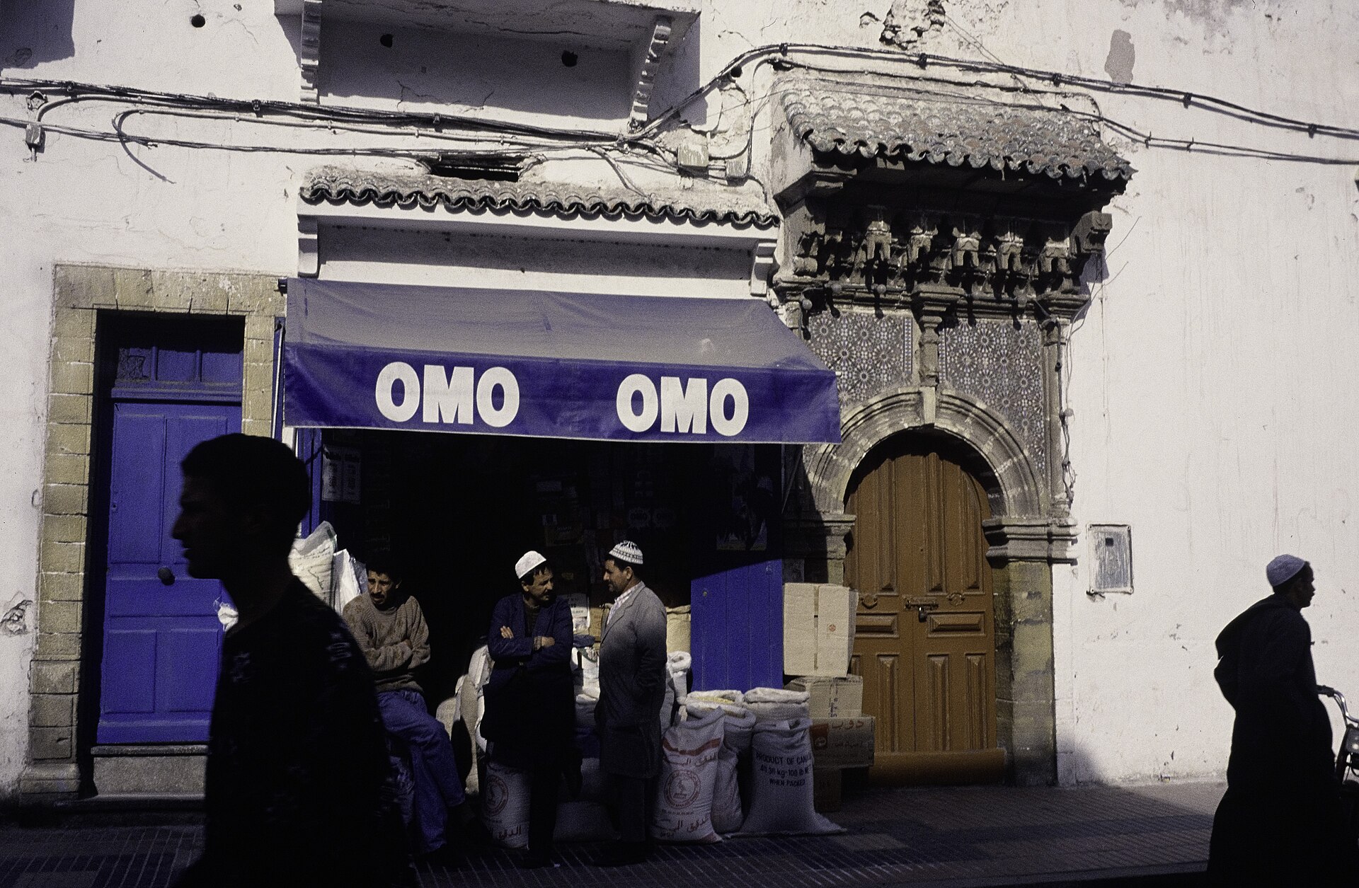 Trois hommes devant une boutique de rue aux sacs de toile de jute, à côté d'une porte décorée, à Essaouira.